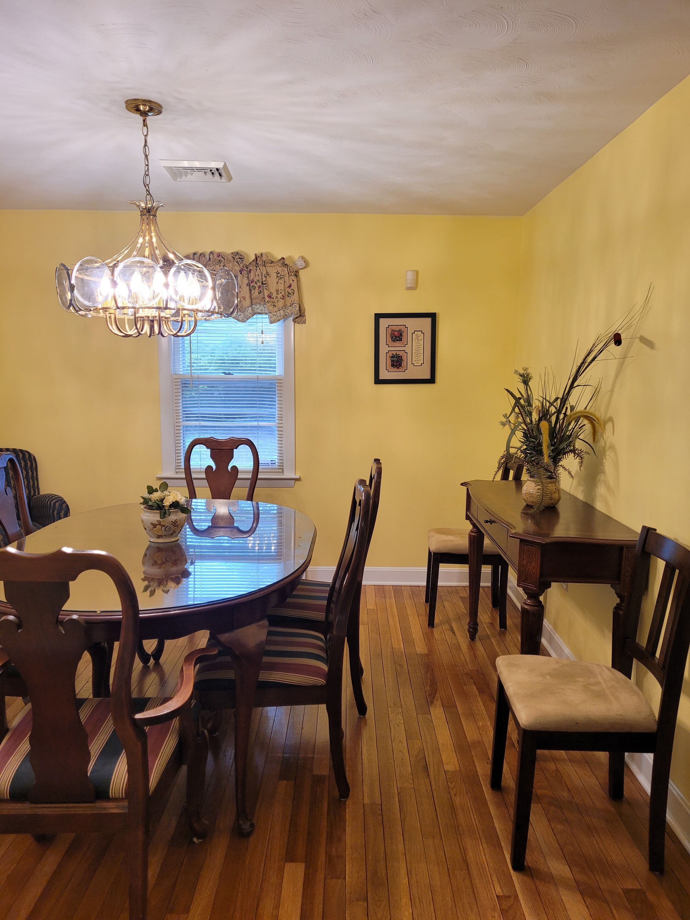 Uma sala de jantar com uma mesa redonda, cadeiras, um lustre, uma janela com persianas, um quadro emoldurado e um vaso de planta.