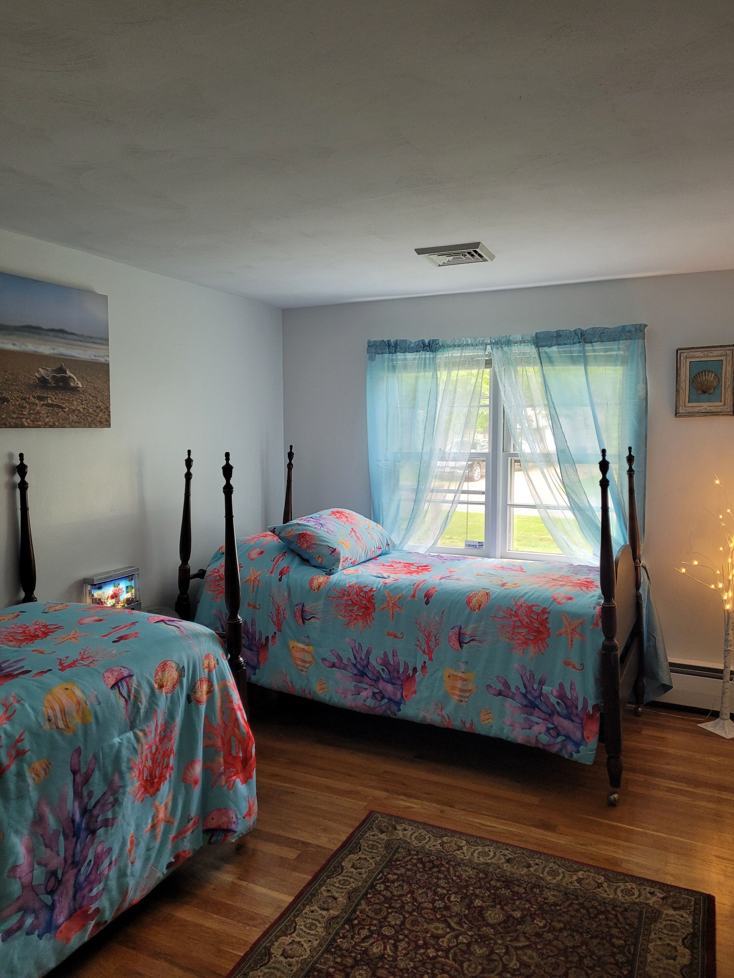 Un dormitorio con dos camas, una mesita de noche, una ventana con cortinas translúcidas, un cuadro enmarcado y una planta decorativa.