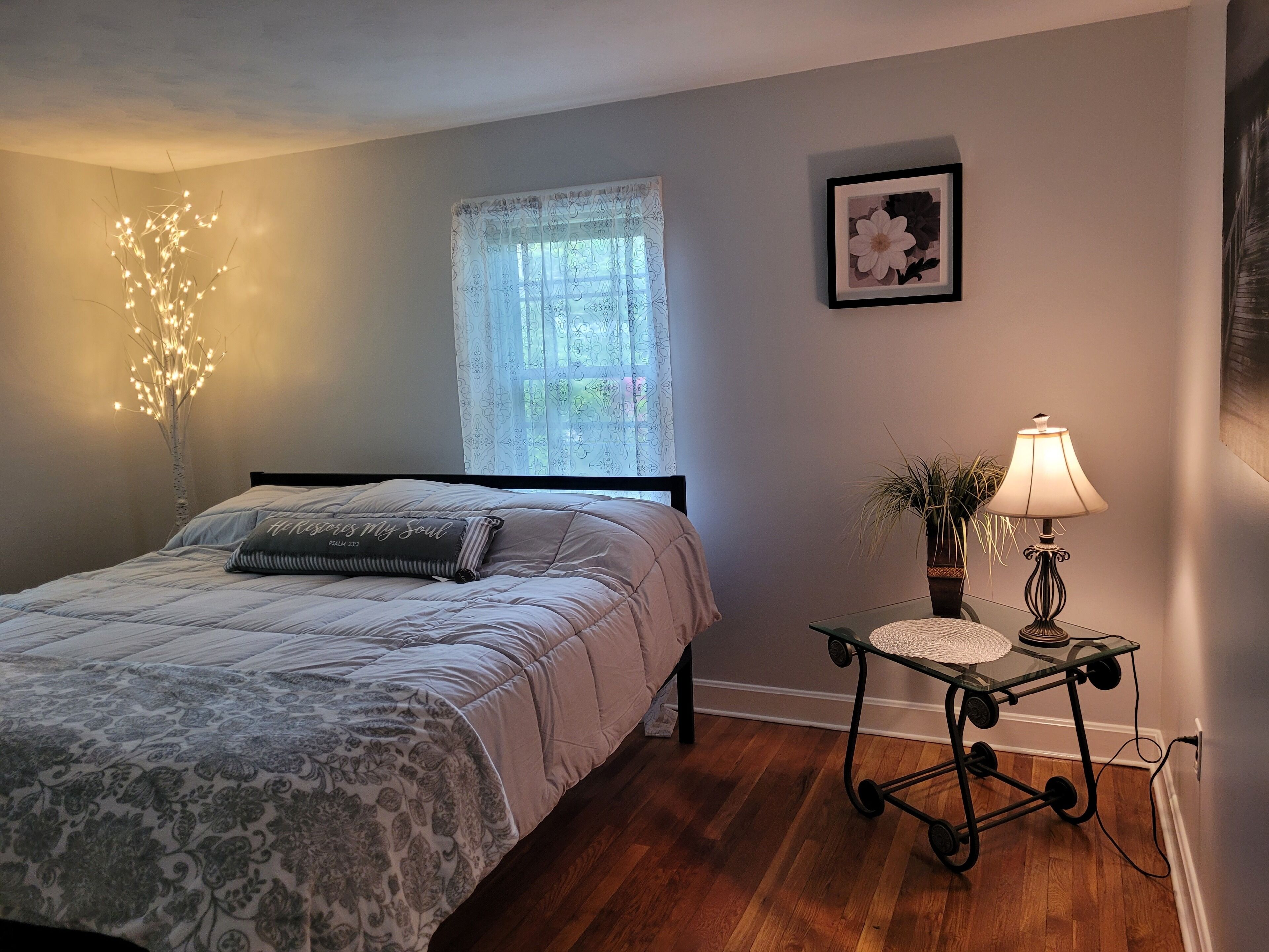 A bedroom with a bed, a bedside table with a lamp, a window with patterned curtains, and a wall-mounted picture.