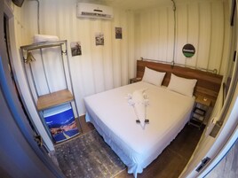 Habitación de hotel con una cama bien hecha, cabecera de madera, aire acondicionado de pared y un estante pequeño con una toalla.