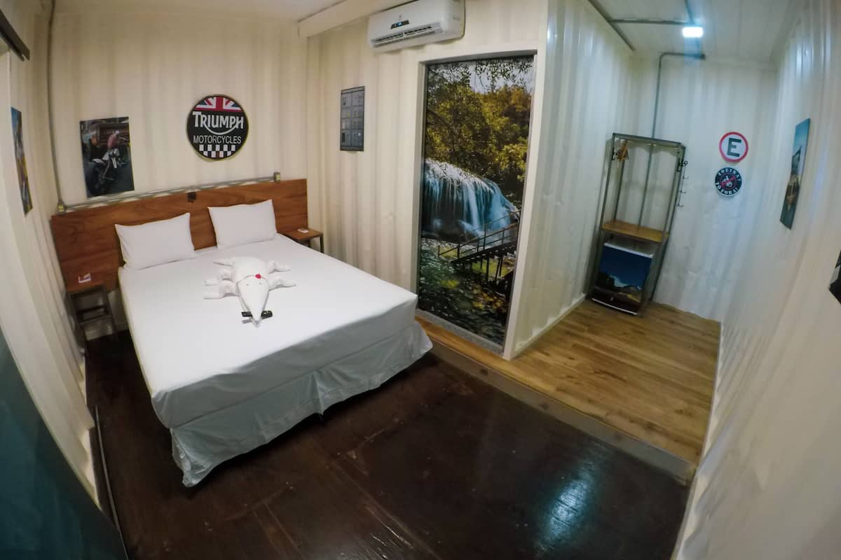 Une chambre d’hôtel avec un lit, une tête de lit en bois, un climatiseur fixé au mur, une affiche de motos Triumph, une affiche d’une cascade et une porte avec un panneau indiquant « accès interdit ».