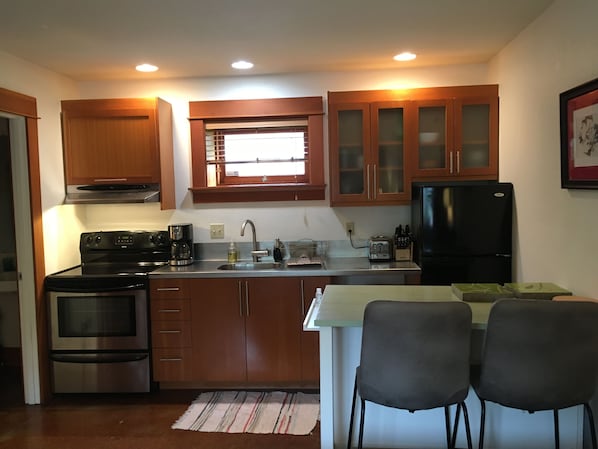 A compact kitchen with wooden cabinets, a stainless steel refrigerator, a built-in oven, and a window above the sink. There are two chairs at a small table.