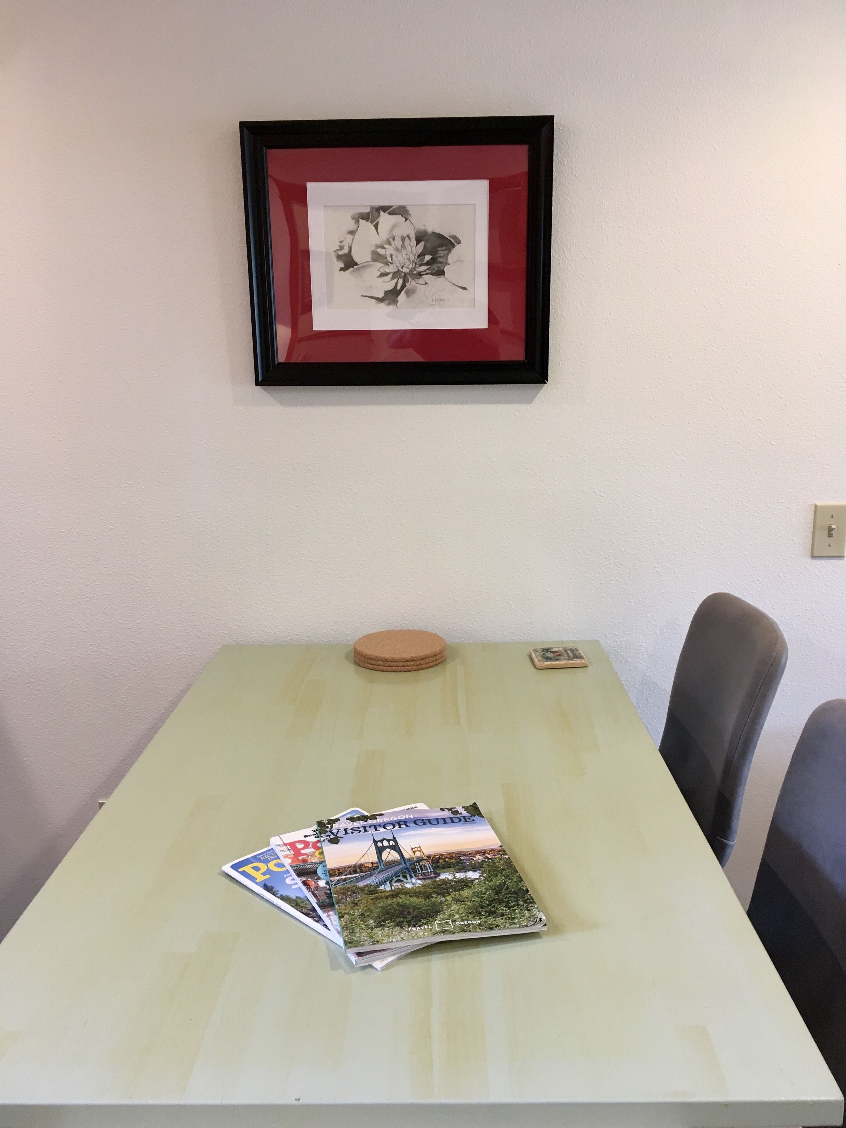 Une table sur laquelle est posée une pile de magazines, avec un tableau encadré au-dessus, et deux chaises à droite.