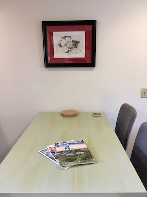 A table with a framed picture above it, a stack of magazines on the table, and two chairs to the right. - Urban Hub, Alberta Arts -ADA (Portland)