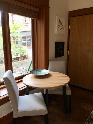 Une pièce comprenant une table ronde en bois, deux chaises blanches avec des coussins, une fenêtre avec des stores, une porte et deux œuvres d’art accrochées au mur.