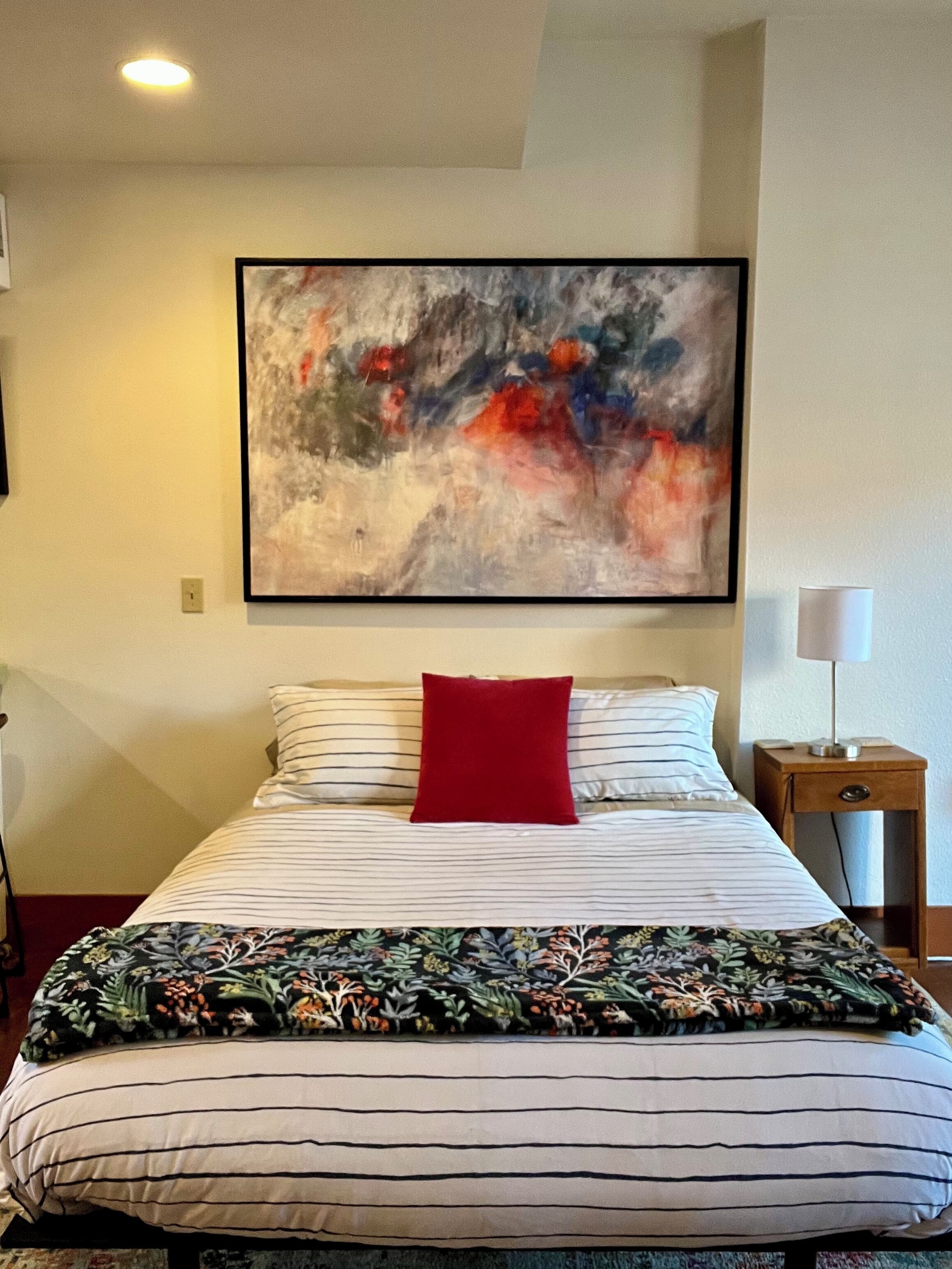 A hotel room with a large bed, a red pillow, a floral bedspread, a nightstand with a lamp, and a painting on the wall.