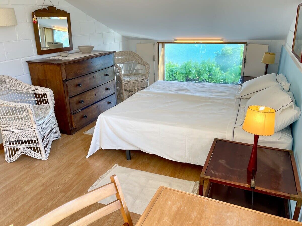 A bedroom with a bed, wooden dresser, wicker chairs, and a window with a view of greenery.