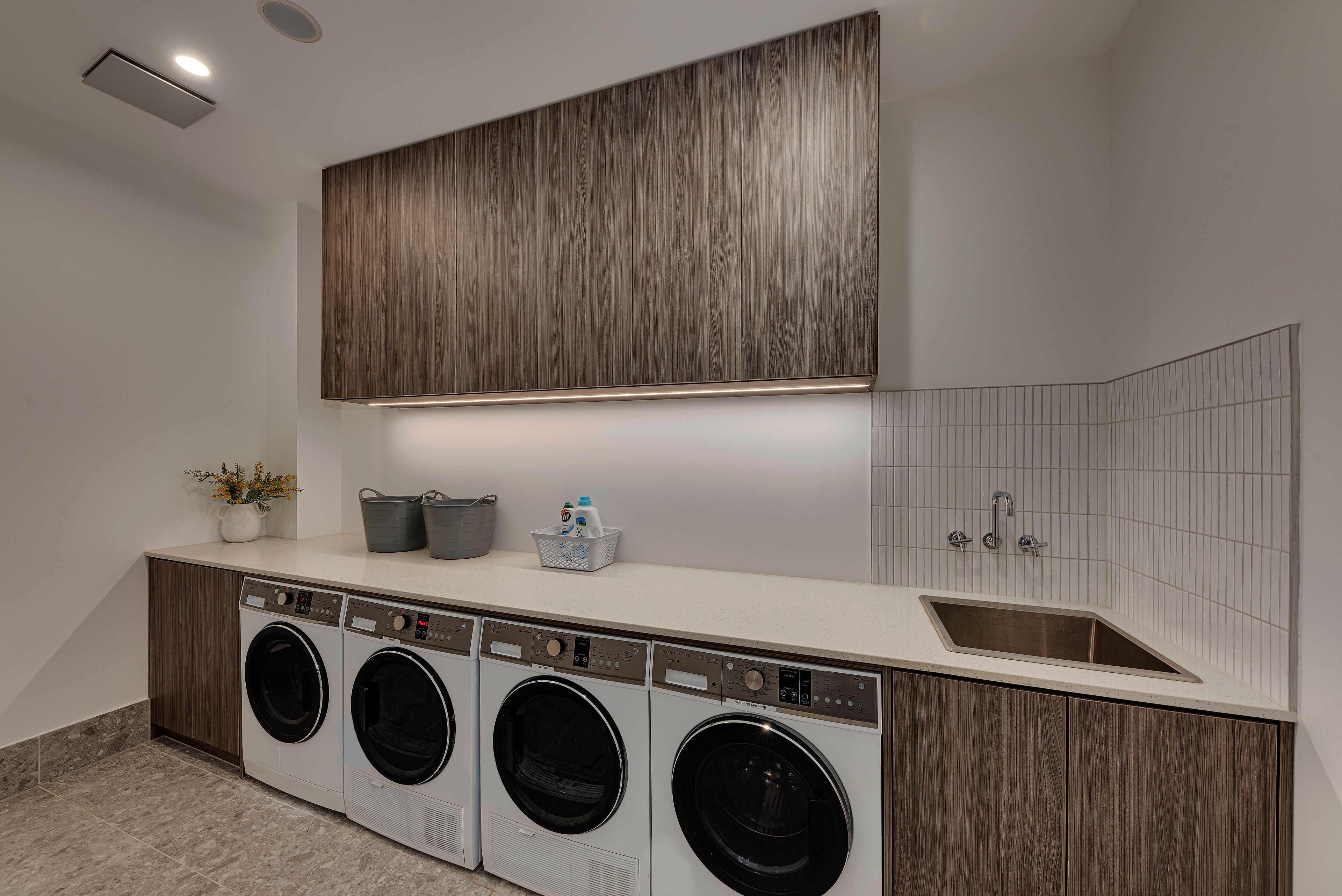 Un moderno lavadero con cuatro lavadoras de carga frontal, una encimera con lavabo y un panel de madera.