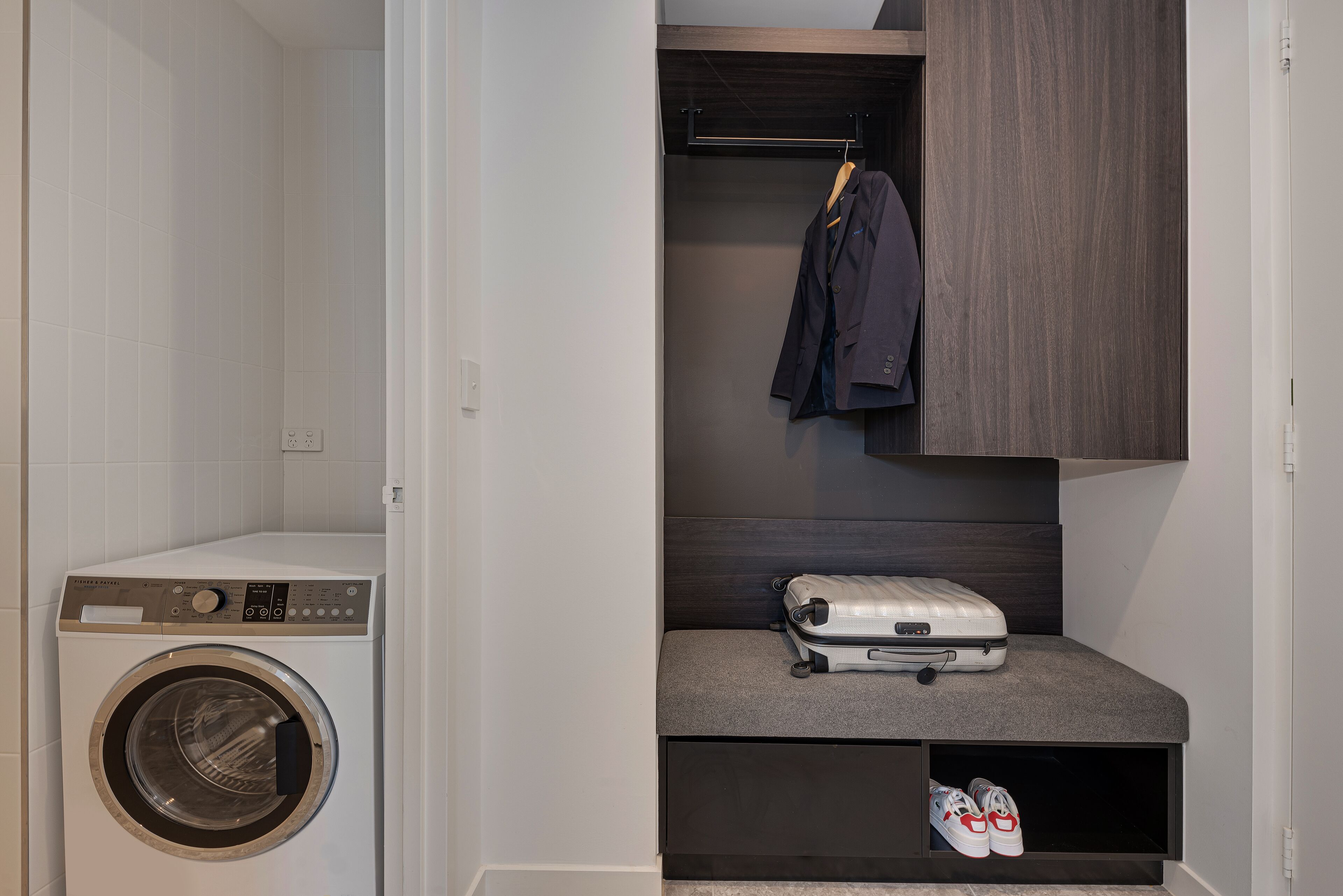 A compact laundry area with a front-loading washing machine, a bench with a suitcase and shoes, and a hanging rack with a jacket.