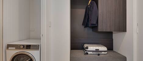 A compact laundry area with a front-loading washing machine, a bench with a suitcase and shoes, and a hanging rack with a jacket.