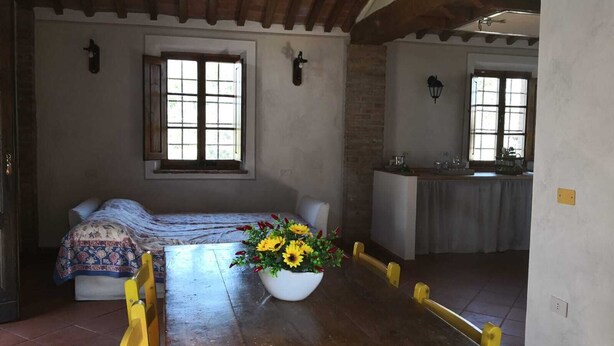 Une pièce rustique avec un banc en bois, une table à manger agrémentée d’un vase de fleurs et, au fond, un coin cuisine.
