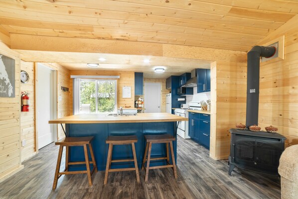 A wooden cabin interior with a kitchen area featuring a central island with stools, a stove, and blue cabinets. - 🏝 Sandy Hooked  2Bed1Bath Remodelled Pine Cottage (Sandy Hook)