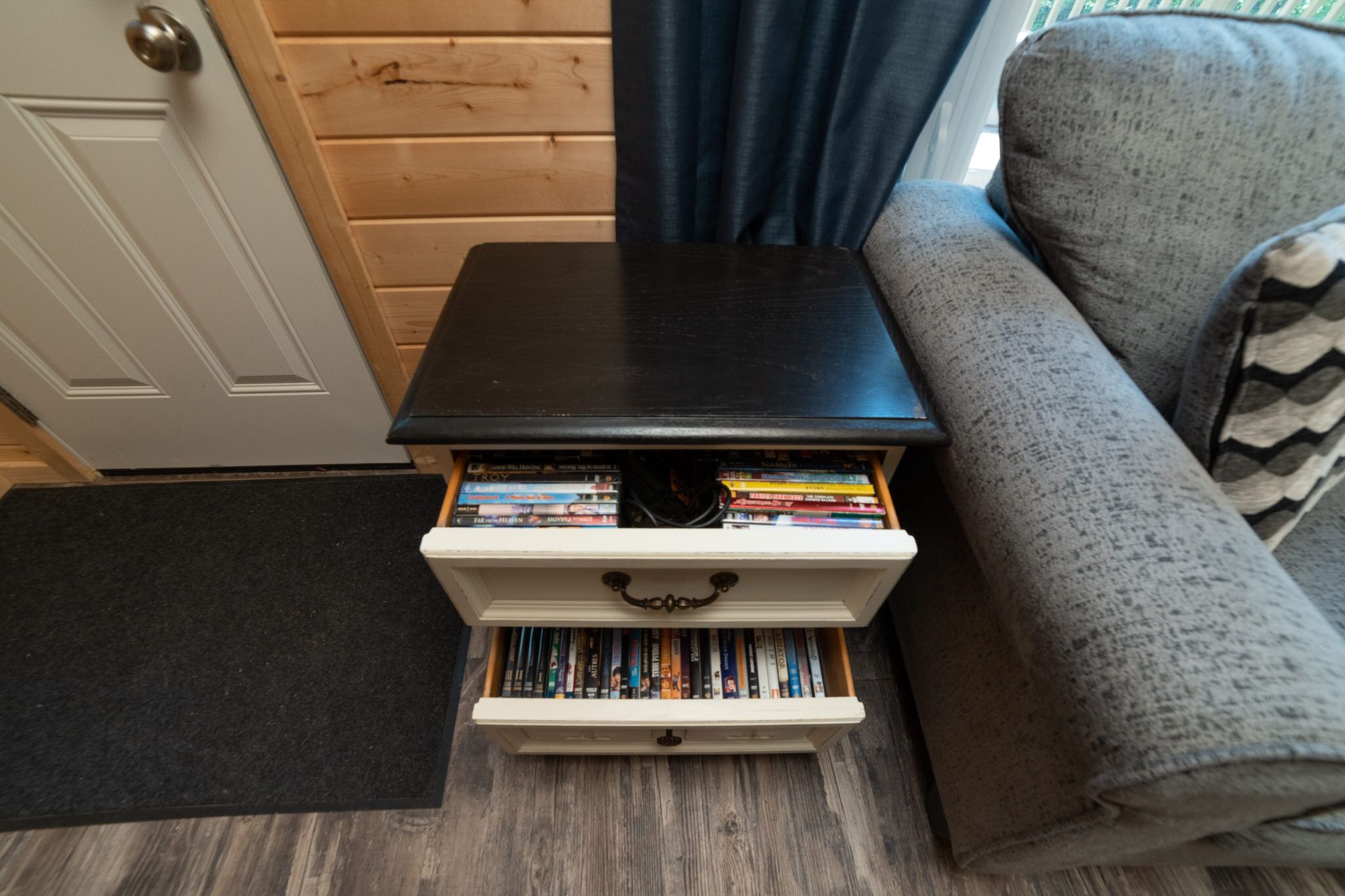 Une petite commode blanche, surmontée d’un plateau noir, remplie de livres et de DVD, est placée à côté d’un canapé gris dans une pièce aux murs en bois et dotée d’un paillasson noir.