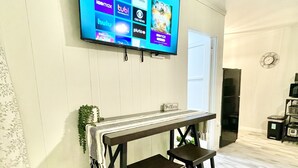 Sala de estar con un televisor de pantalla plana montado que muestra varios servicios de streaming, una mesa de madera con dos taburetes y una planta en maceta.