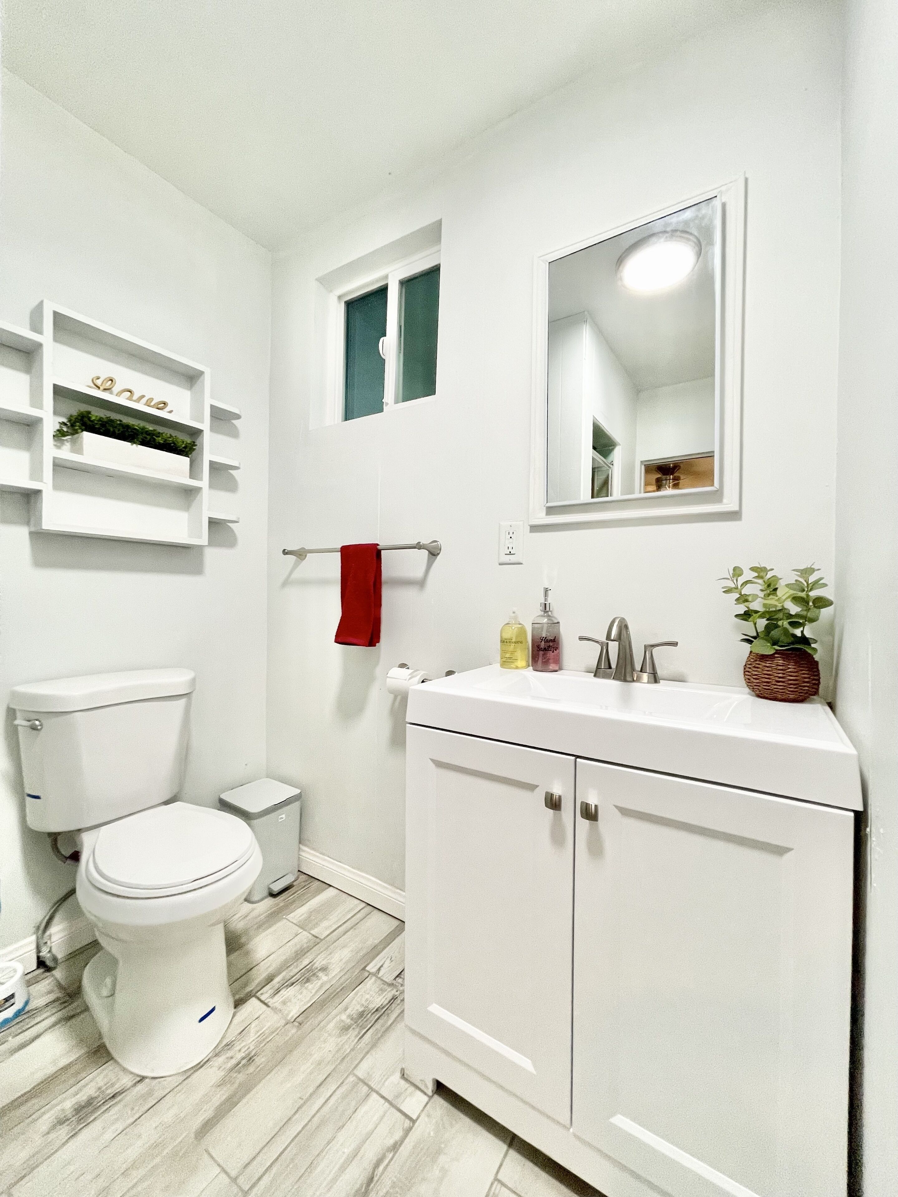 Un baño pequeño y blanco con inodoro, lavabo y espejo. Hay un estante sobre el inodoro con una planta y un cartel.