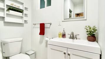 Un baño pequeño y blanco con inodoro, lavabo y espejo. Hay un estante sobre el inodoro con una planta y un cartel.