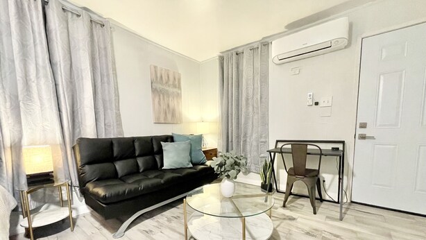 Una sala de estar moderna con un sofá de cuero negro, una mesa de centro de vidrio, una mesa de comedor con una silla y una puerta blanca.