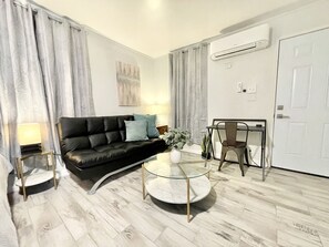 A modern living room with a black leather sofa, a glass coffee table, a dining table with a chair, and a white door.
