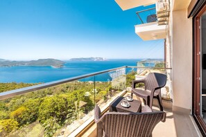 Un balcón con muebles de mimbre, una mesa con tazas y una vista panorámica del mar y las colinas.