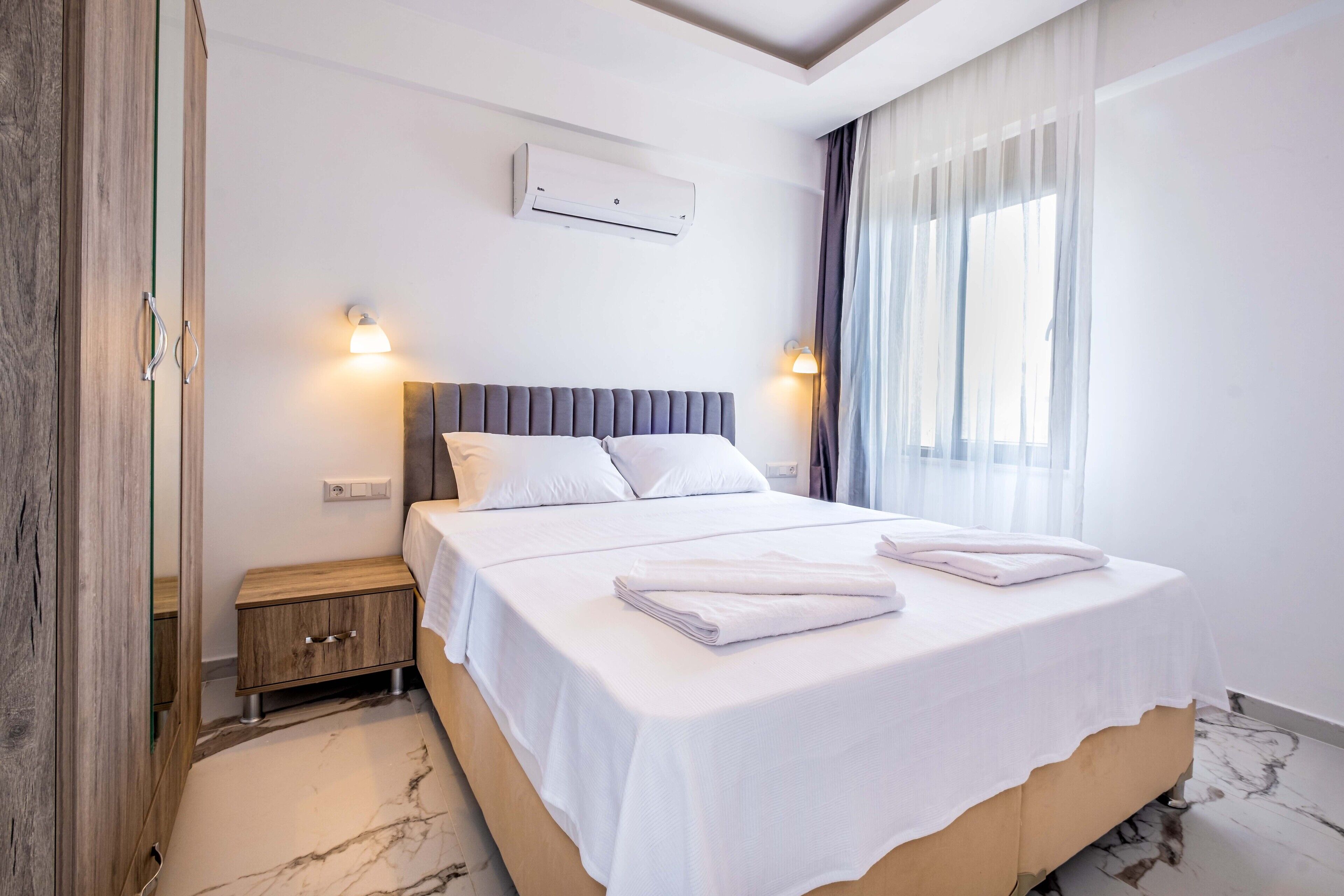 Ein Hotelzimmer mit einem großen Bett, einem Nachttisch aus Holz, einer Wand-Klimaanlage und einem Fenster mit durchsichtigen Vorhängen.