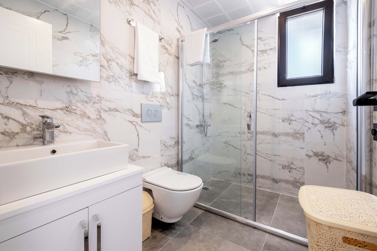 A modern bathroom with a marble backsplash, a glass shower enclosure, and a white sink.