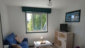 A room with a blue sofa, a wooden cabinet with a TV, a window with a view of trees, and a small table with a flower. - Apartment close to the beach (Houlgate)