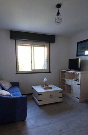 A living room with a blue sofa, a white coffee table, a television on a wooden stand, and a window with blinds.