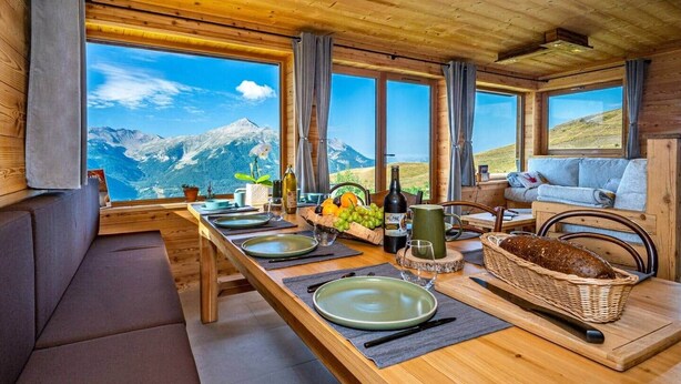 Ein Holzespeisetisch ist mit Tellern, Besteck und einer Weinflasche gedeckt, mit Blick auf Berge durch große Fenster.