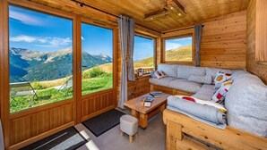 Ein gemütliches Blockhaus mit Holzeinrichtung, großen Fenstern mit Blick auf die Berge, einer Couch und einem kleinen Tisch.