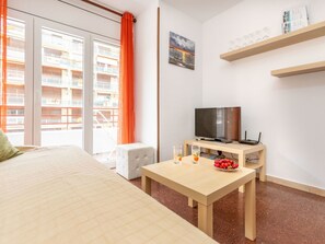 A hotel room with a bed, a TV, a small table with drinks and fruit, a window with curtains, and a balcony with a view of buildings.