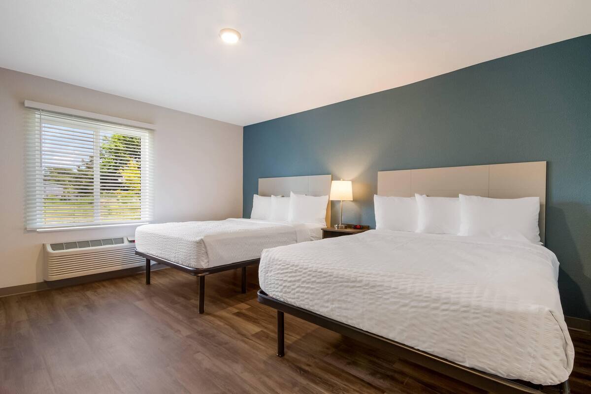 Ein Hotelzimmer mit zwei Betten, einem Fenster mit Jalousien, einem Heizgerät und einem Nachttisch mit Lampe.
