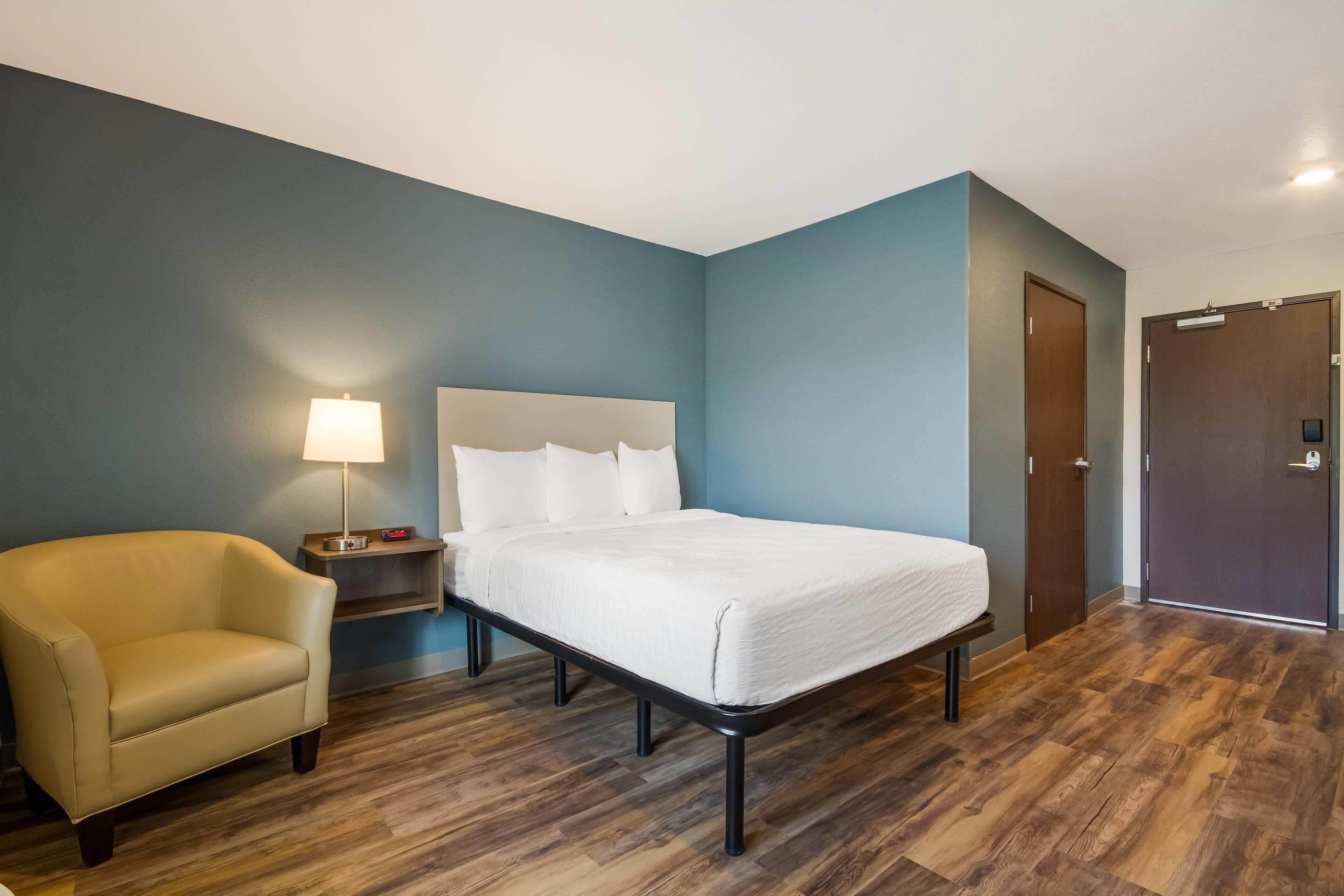 Una camera d'albergo con un letto, una sedia, un comodino con una lampada e una porta.