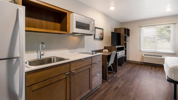 A compact kitchen with a refrigerator, microwave, and sink. There is a chair and a small table with a chair. The room has wooden flooring and a window with blinds. - Woodspring Suites Panama City Beach (Panama City Beach)