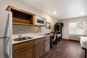 A compact kitchen with a refrigerator, microwave, and sink. There is a chair and a small table with a chair. The room has wooden flooring and a window with blinds. - Woodspring Suites Panama City Beach (Panama City Beach)