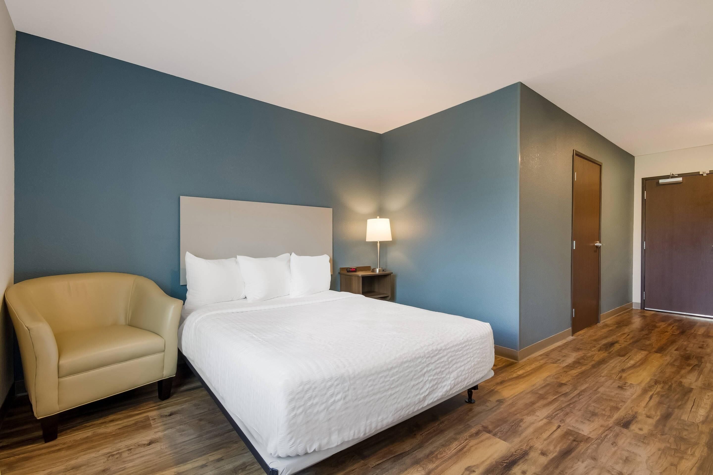Una habitación de hotel con una cama, una silla, una mesita de noche con lámpara y una puerta.