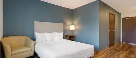 Una habitación de hotel con una cama, una silla, una mesita de noche con lámpara y una puerta.