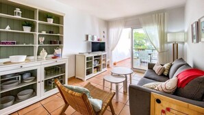 A living room with a sofa, a TV, and a dining area with a table and chairs. There is a bookshelf with various items and a window with curtains. - Golf Royal Omega by Interhome (Pals)