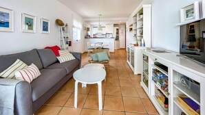 A compact living space with a grey sofa, a small white table, a kitchen area with white cabinets, and a dining table with chairs. - Golf Royal Omega by Interhome (Pals)