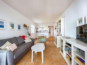 A compact living space with a grey sofa, a small white table, a kitchen area with white cabinets, and a dining table with chairs.