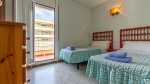 A room with two beds, a small table, a wooden dresser, and a balcony with a view of buildings. - Atlantis by Interhome (Segur de Calafell)