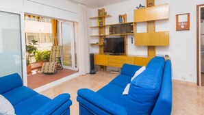 A living room with a blue sofa set, a TV on a wooden stand, and a sliding glass door leading to an outdoor area with plants. - Atlantis by Interhome (Segur de Calafell)
