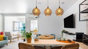 A modern kitchen with a dining area, featuring a round table, wicker chairs, hanging pendant lights, and a flat-screen TV mounted on the wall.