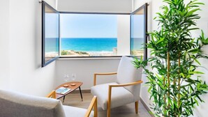 A modern room with a large window offering a view of the sea. There is a comfortable chair, a small table with glasses, and a tall green plant.