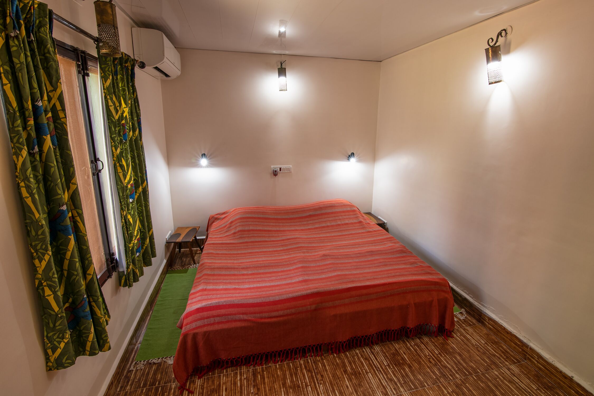 A bedroom with a bed, green patterned curtains, a red blanket, and two wall-mounted lights.