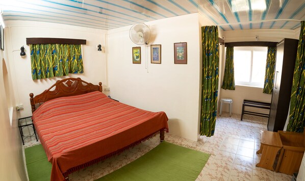 A bedroom with a bed, a standing fan, a window with curtains, and a small stool. - Bamboo banks (Ootacamund)