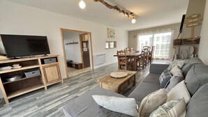 A modern living room with a grey sofa, wooden coffee table, and a flat-screen TV on a wooden stand. There is a dining area with a wooden table and chairs, and a large window with curtains. - House (Asnelles)