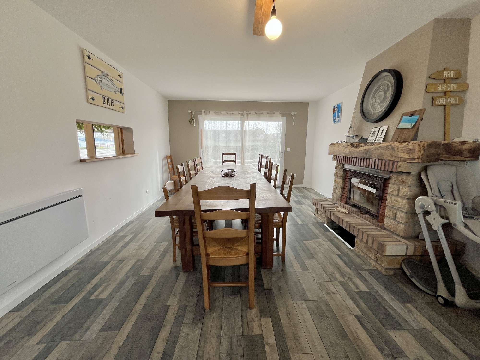 A dining area with a long wooden table and chairs, a fireplace, and a view through large windows.