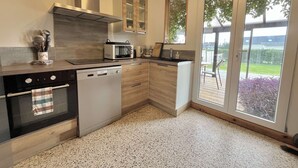 A modern kitchen with a dishwasher, microwave, and stove, wooden cabinets, and a sliding glass door to a deck. - House (Asnelles)