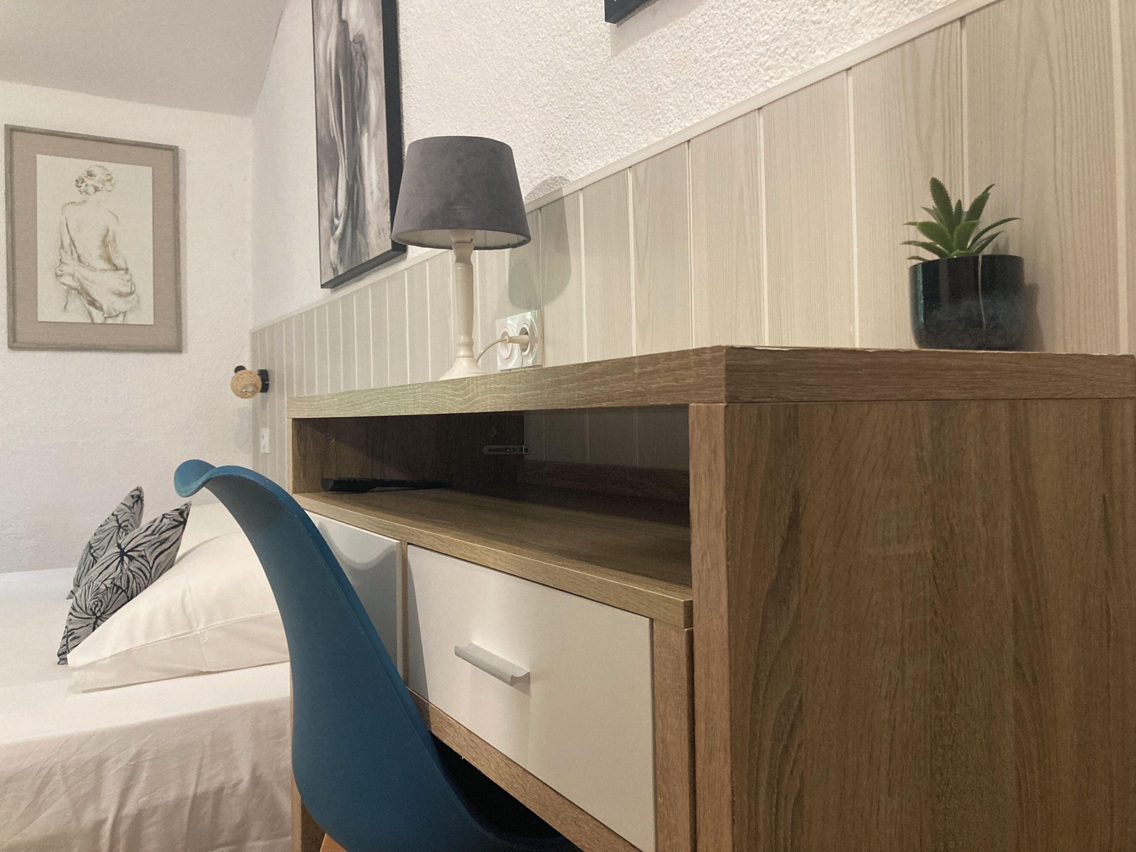 Une chambre à coucher avec un lit, un bureau, une chaise, une lampe, une plante et un tableau accroché au mur.