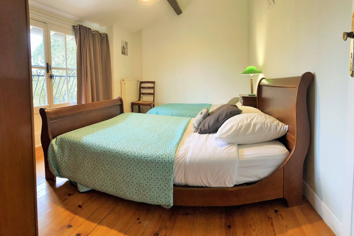 Une chambre à coucher avec un lit en bois, une chaise, une table de chevet, une lampe et une fenêtre avec des rideaux.